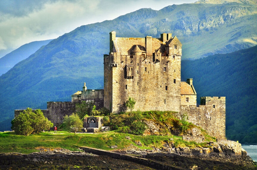 A stone citadel stands on a green hill, surrounded by mountains under a cloudy sky. A person in a red jacket walks on a path near the castle.