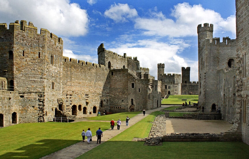 A group of people walk along a pathway surrounded by ancient stone walls and towers at a historic citadel on a partly cloudy day.