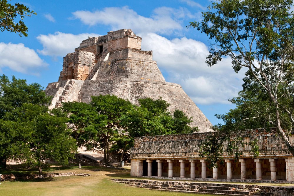 Ancient stone pyramid with steep steps surrounded by trees and smaller stone structures under a clear blue sky, unlocking the secrets of the Maya civilization.