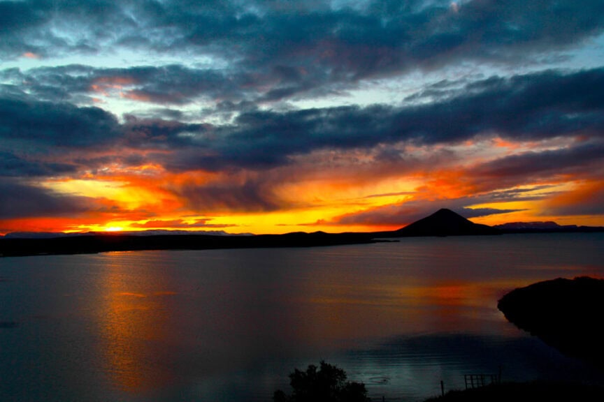 Sunset over a calm lake with vibrant orange and blue hues in the sky, a silhouetted mountain in the background, and dark clouds adding dramatic contrast, reminiscent of a painter's palette.