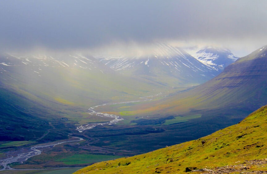 A valley with a winding river runs through green fields and snow-capped mountains under a cloudy sky, as if nature itself provided the perfect painter's palette.