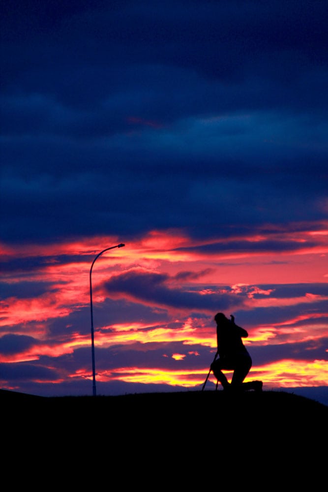 A silhouetted person kneels near a streetlight, their hand clutching painting tools, against a vibrant sunset sky with dark clouds.