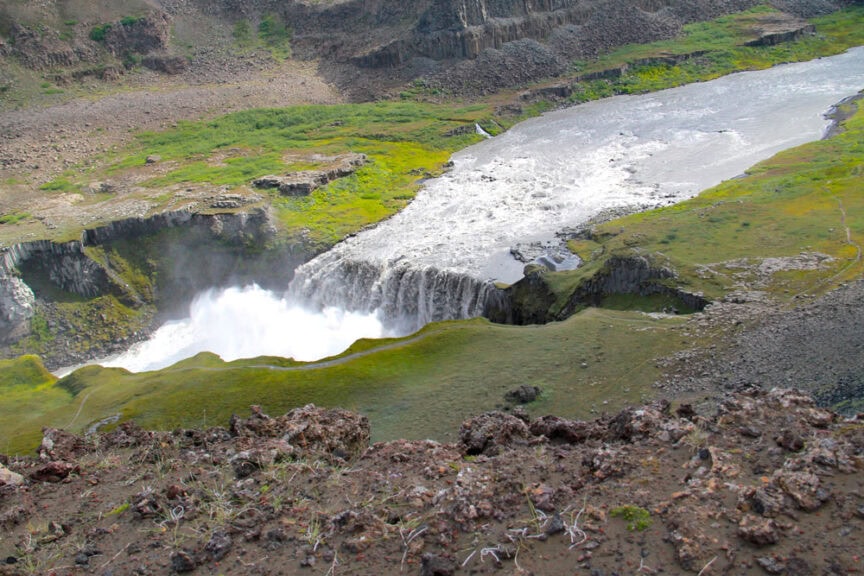 A waterfall flows over rocky terrain into a misty plunge pool below, with a green and rocky landscape resembling a painter's palette.