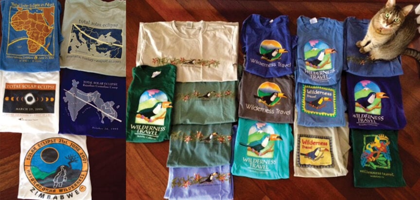 An arrangement of various t-shirts featuring wilderness and eclipse designs, with a cat sitting beside the collection on the right.