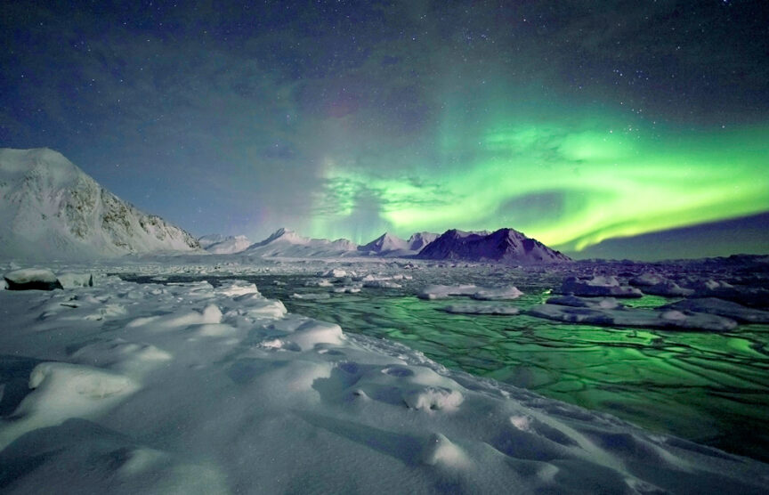 A 2016 adventure in a snow-covered landscape reveals a partially frozen lake and mountain range under a night sky illuminated by green and yellow auroras.