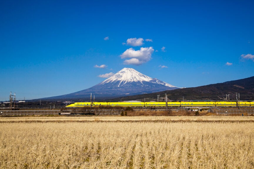 A fun yellow train speeds through fields with snow-capped Mount Fuji in the background, under Japan's clear blue sky.