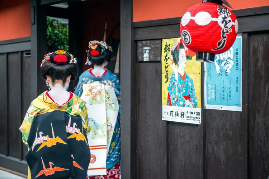 Two women in traditional Japanese kimono attire, with intricate hair accessories, stand near a wooden building entrance decorated with a red lantern and posters. Fun fact: such scenes are common during festivals in Japan, showcasing the country's rich cultural heritage.