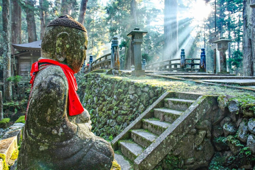 A stone Buddha statue with a red cloth stands near stone steps and a bridge in a sunlit forest with rays filtering through trees, evoking the serene landscapes of Japan.