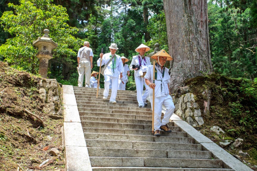 A group of people dressed in white attire and traditional hats descend a stone staircase in a forested area, reminiscent of cultural customs often seen in Japan.
