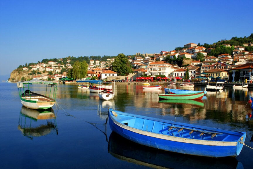 A serene lakeside scene with several colorful boats floating on calm water and a village with houses and buildings on a hillside in the background captures the Spirit of Albania.