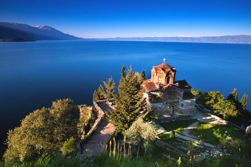 The stone church with its red roof, surrounded by trees and overlooking a large, serene lake with mountains in the distance under a clear blue sky, captures the Spirit of Albania.