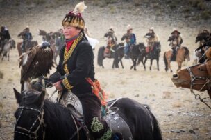 A person dressed in traditional clothing rides a black horse while holding a golden eagle, showcasing their skills at the Golden Eagle Festival. A group of similarly dressed individuals on horseback are seen in the background, adding to the event's vibrant atmosphere.