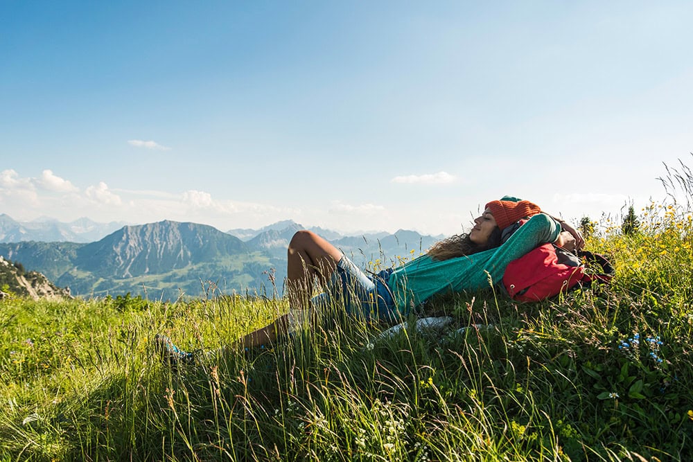 Person lying on a grassy hill with a scenic mountain view in the background, wearing a green sweater and an orange hat, with a red backpack by their side, embracing secrets of healthy living.