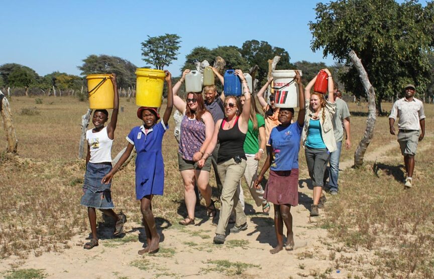 A group of people, including locals and visitors, carry water buckets on their heads while walking along a dirt path in a rural area. Trees and open fields are visible in the background, showcasing a travel experience that gives back to the community.