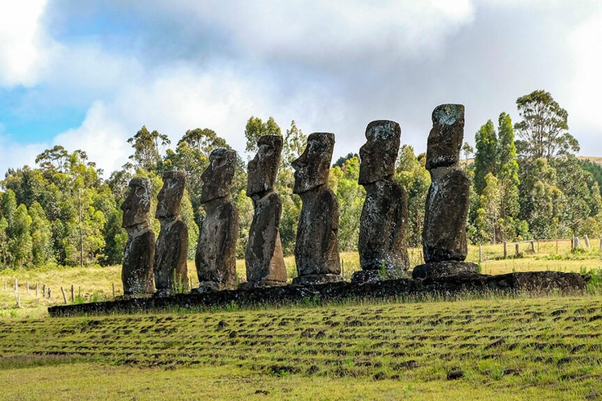 Several moai statues stand in a row on a grassy hill, shrouded in the mysteries of Tangata Manu, with trees and a cloudy sky in the background.