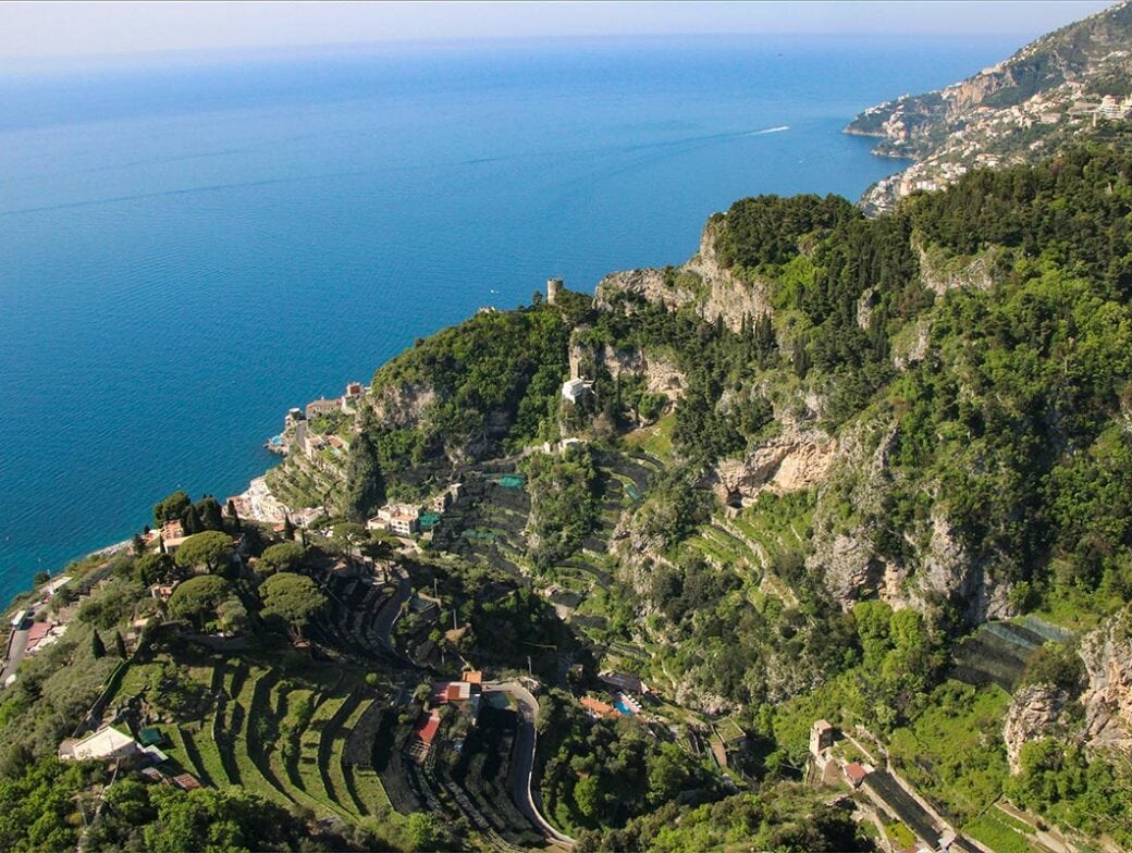 Aerial view of hilly coastal landscape with terraced greenery and scattered buildings along the coastline of a clear blue sea, reminiscent of the stunning Amalfi coast and perfect for summer adventures.