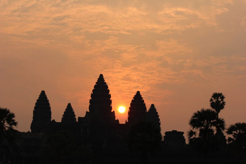 Silhouette of Angkor Wat temple with the sun setting behind its towers, under a cloudy orange sky, framed by palm trees.