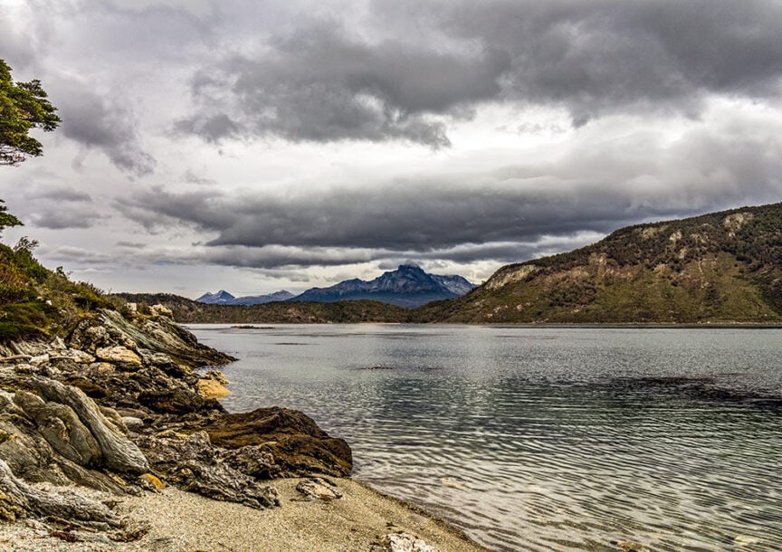 A serene landscape of a rocky shore leading to a calm body of water, with distant mountains under a cloudy sky, evokes the tranquil beauty found at the End of the World in Ushuaia.