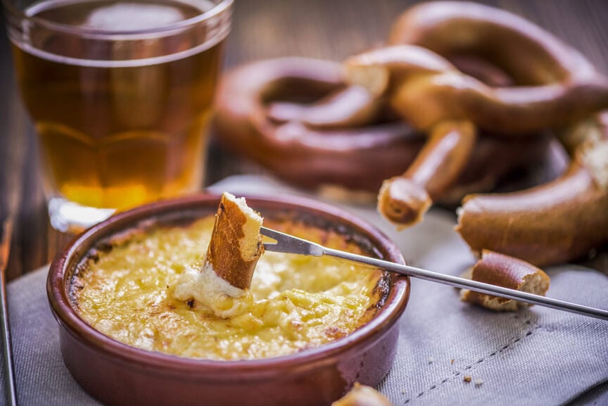 A piece of bread is being dipped into a cheese fondue in a brown dish, reminiscent of an après-hike treat in the Alps, with a glass of beer and pretzels in the background.