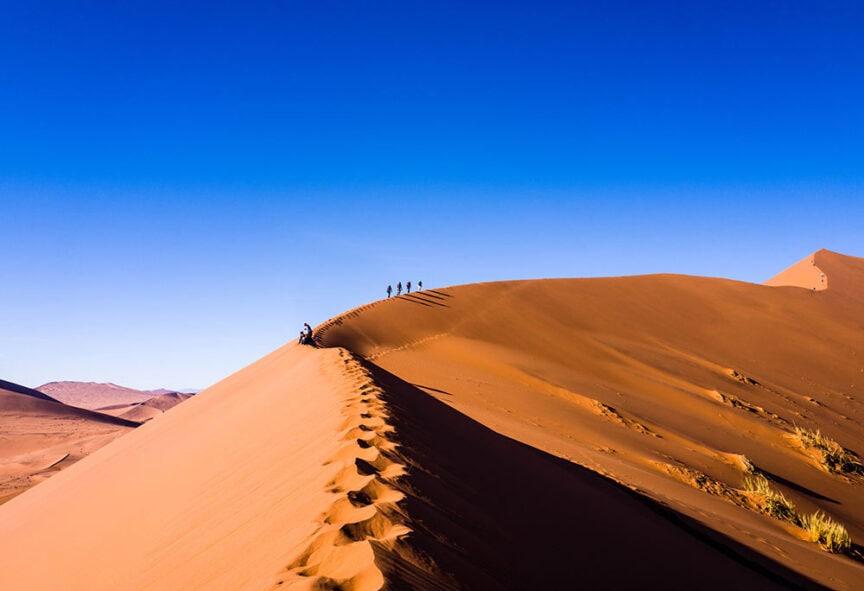Five people walking on the ridge of a large sand dune in Namibia under a clear blue sky. Footprints trail behind them on the orange sand, painting a mesmerizing landscape against the vast horizon.