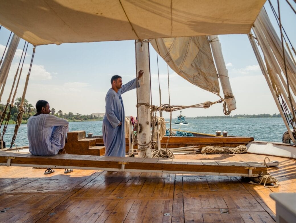 Two men in traditional clothing are on a wooden sailboat, or Dahabiya, gently gliding along the calm waters of the Nile River, with one man sitting and the other standing by the sail. Palms and distant land enhance their serene exploration.