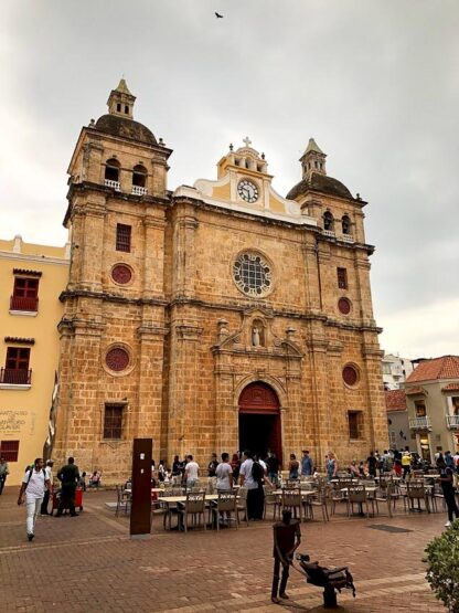 People gather in front of a large, historic church with two bell towers and a clock in the center facade. The church is surrounded by a plaza with tables and chairs, offering amazing experiences as part of Colombia travel. The sky is overcast.