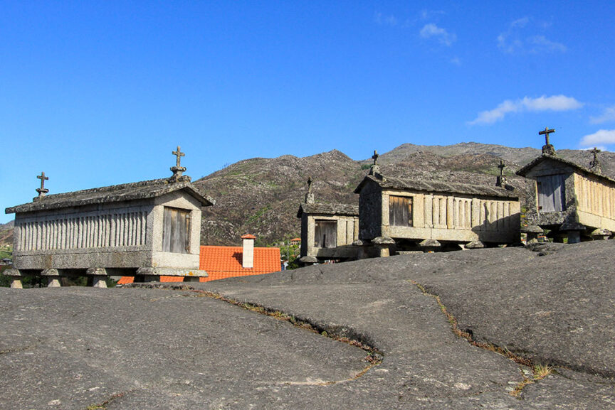 Several stone granaries with cross-topped roofs sit elevated on stilts in a rocky landscape against a mountainous backdrop, capturing the simple pleasures of rural Portugal.
