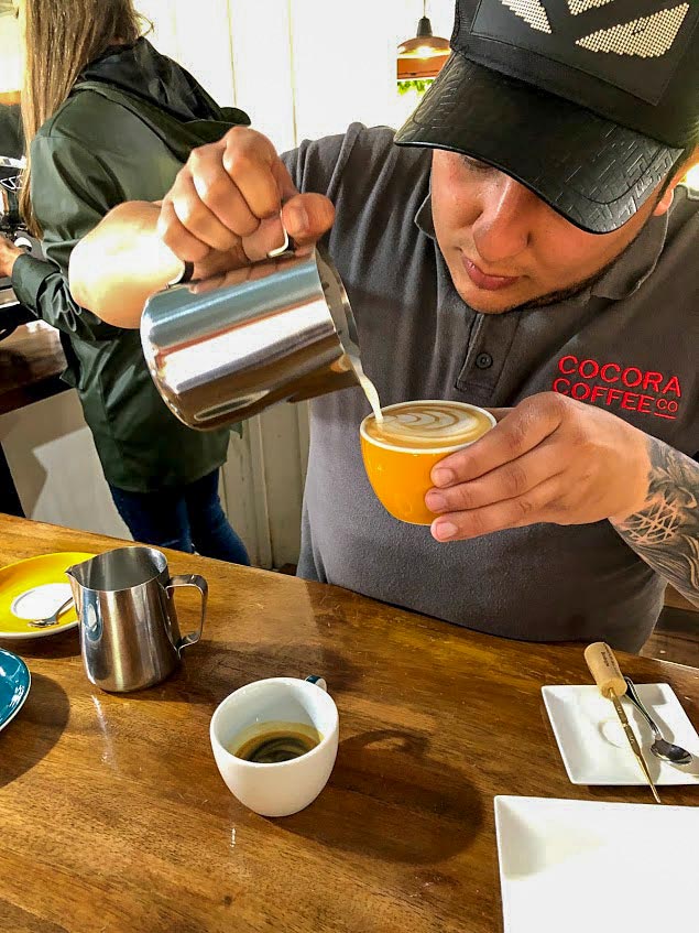 A barista wearing a cap and a shirt with "Cocora Coffee Co" logo pours milk into a cup of coffee on a wooden counter, delivering amazing experiences. An espresso shot and a milk frothing pitcher are also on the counter, highlighting the rich coffee traditions of Colombia.