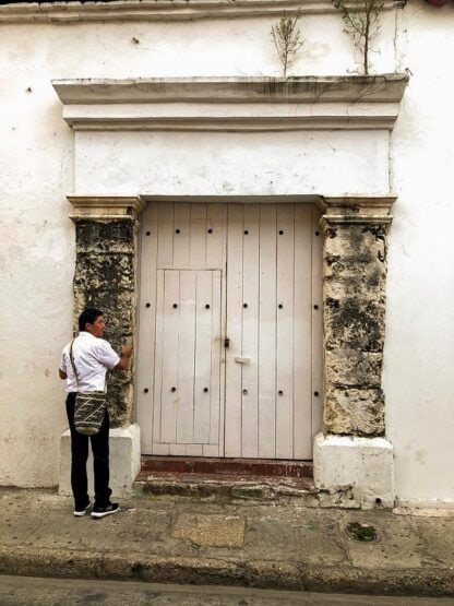 A person stands in front of a large, weathered wooden door set within a stone frame, knocking or attempting to open it. The person is wearing a white shirt and carrying a black and white patterned bag, ready for amazing experiences in Colombia.