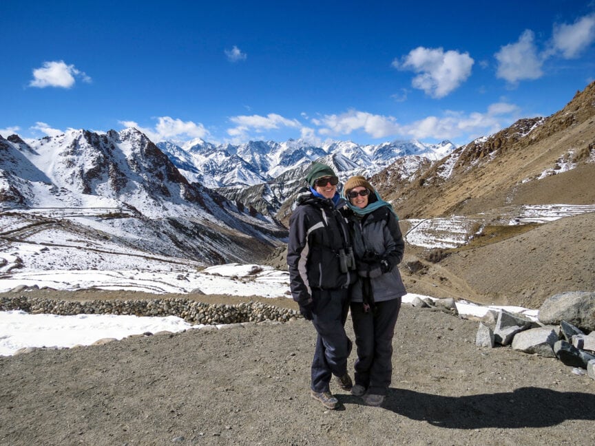 Two people in winter clothing stand together on a rocky path, with snow-capped mountains reminiscent of India's majestic ranges and a clear blue sky in the background, conjuring visions of elusive Snow Leopards.