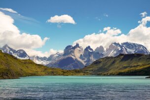 A turquoise lake nestles into the green hills and rugged, snow-capped mountains of Patagonia, all under a partly cloudy blue sky.