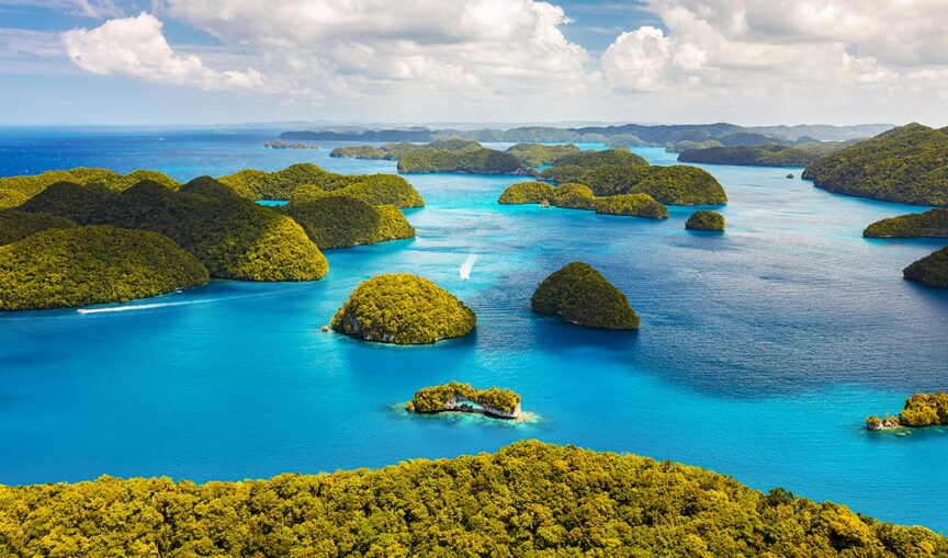 Aerial view of numerous small, lush green islands scattered in vibrant blue water under a partly cloudy sky. A boat, resembling an adventure short film scene, leaves a white trail in the middle distance.