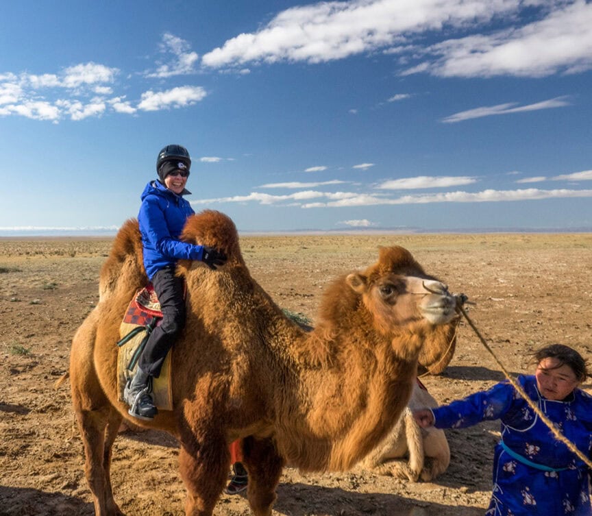 A person wearing a blue jacket and helmet sits on a camel in the Gobi Desert while another individual in traditional attire, reminiscent of the rich cultural heritage of Mongolia, holds the camel's reins.