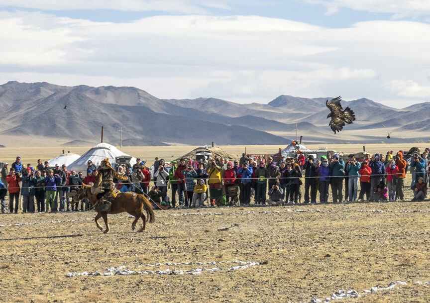 A person on horseback, accompanied by a majestic Golden Eagle, rides past a crowd of spectators in the open, mountainous landscape of Mongolia.