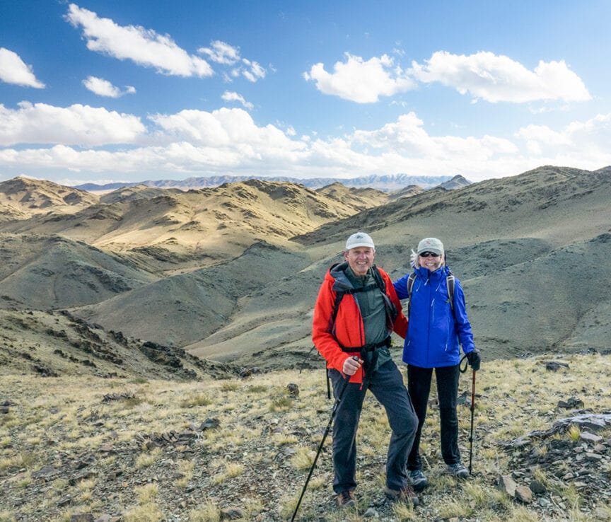 Two hikers, one in a red jacket and one in a blue jacket, stand on a rocky hillside with trekking poles, reminiscent of adventurers exploring the vast landscapes of Mongolia. Behind them, a majestic mountainous expanse stretches under a blue sky dotted with scattered clouds.