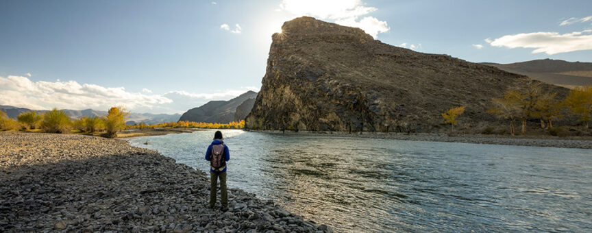 A person with a backpack stands on a rocky riverbank, looking at a large rock formation across the water under a clear sky, reminiscent of the rugged landscapes found in Mongolia's Gobi Desert.