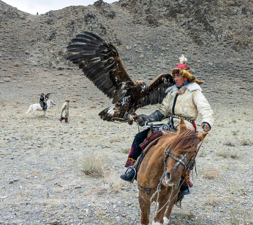 A person rides a horse in a rocky landscape, holding a large golden eagle on their arm. Another rider on horseback is in the background, further emphasizing the remote, mountainous environment of Mongolia.