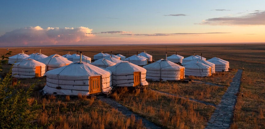 A group of white, circular yurts sits on a grassy plain at sunset in Mongolia, with a stone path leading through the area. In the distance, Golden Eagles soar above this tranquil scene reminiscent of the vast Gobi Desert.