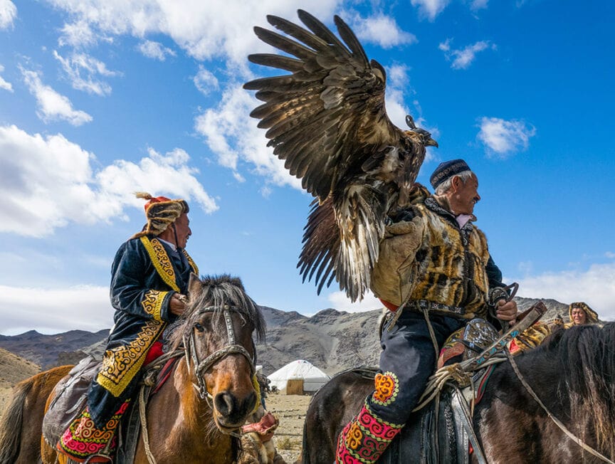 Two individuals in traditional attire on horseback, one of them holding a large golden eagle with its wings spread. A yurt and the vast expanse of Mongolia's mountainous landscape are visible in the background.
