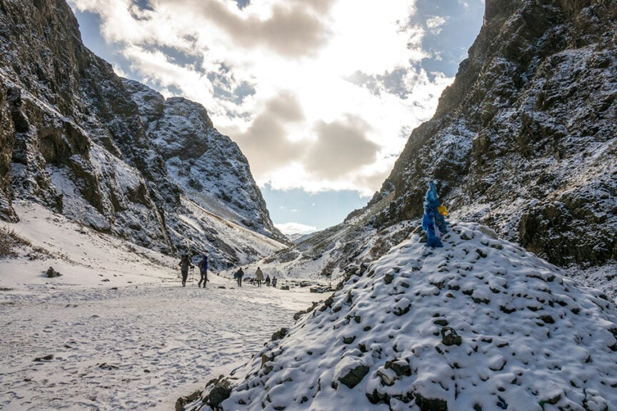 Snow-covered mountainous landscape with people hiking in the distance. A cairn is in the foreground, topped with blue and yellow fabric, reminiscent of tradition found in Mongolia. Clouds are scattered across the bright sky, perhaps offering a glimpse into a terrain where Golden Eagles might soar.