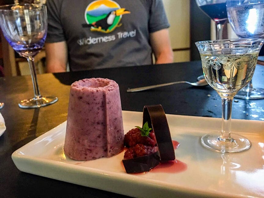A cylindrical berry dessert and chocolate garnish on a plate, accompanied by a glass of white wine. A person wearing an "Epic Hikes" shirt is visible in the background.