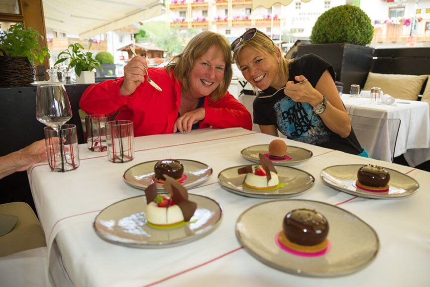 Two women smile and hold forks over plates of various desserts on a table set outside under a canopy, enjoying a sweet break after exploring the Great Dolomites.