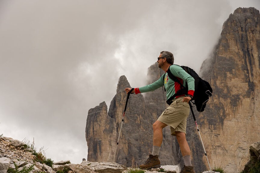 A person stands on rocky terrain with hiking poles, wearing a backpack and exploring the mountainous landscape of the Great Dolomites, enveloped in clouds.