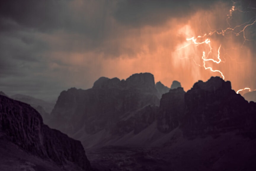 Exploring Dolomites at dusk reveals a mountainous landscape with dramatic clouds and lightning illuminating the dark silhouette of the peaks.