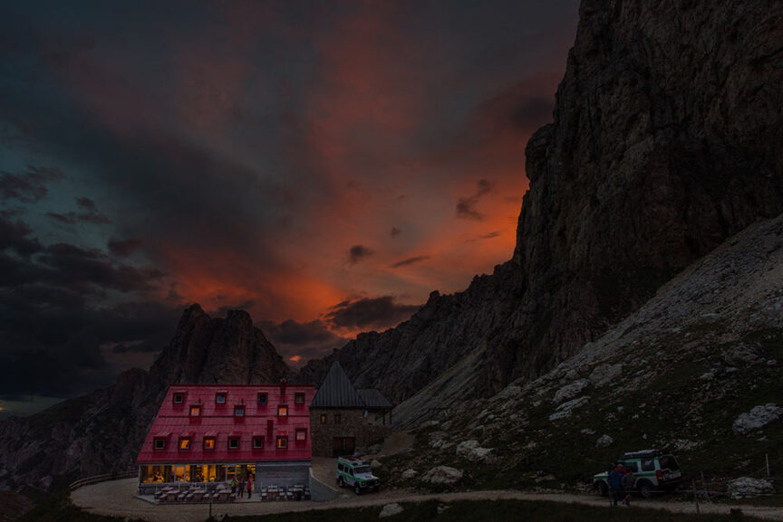 A mountain hut with a bright red roof is set against a dramatic sunset sky with towering rocky cliffs and rugged terrain in the background. Some lights are on in the hut, and a few vehicles are parked nearby, perfect for those exploring the Great Dolomites.