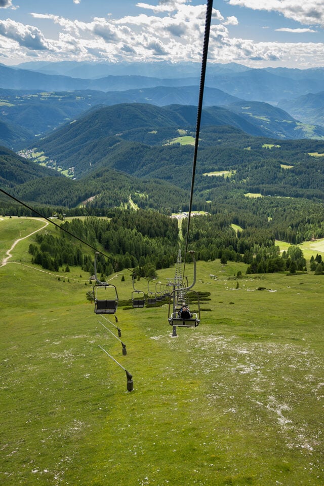 A chairlift ascends a grassy green mountain with a backdrop of tree-covered hills and a cloudy sky, offering an ideal vantage point for exploring the beauty of the Great Dolomites.