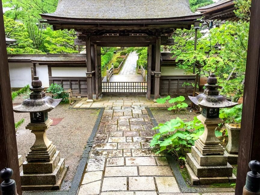 A traditional Japanese stone pathway leads through a wooden gate, flanked by stone lanterns amidst lush green foliage and a serene garden setting, capturing the essence of ancient Japanese culture.