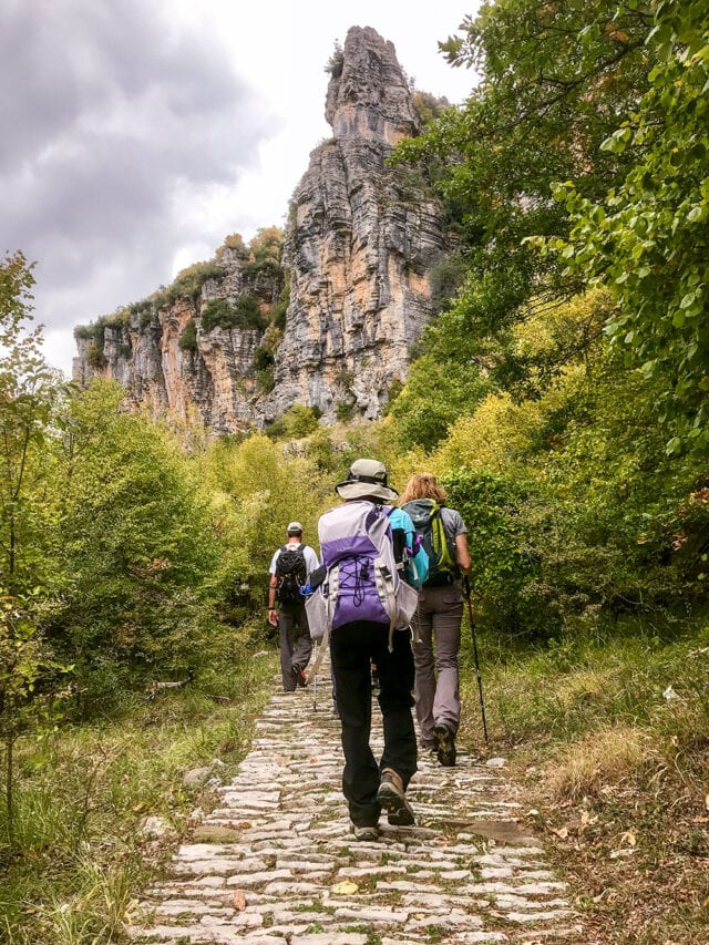 Three people hike on a cobblestone path towards a tall, rocky cliff surrounded by lush greenery under a cloudy sky, reminiscent of the rugged beauty found in the isles of Greece.