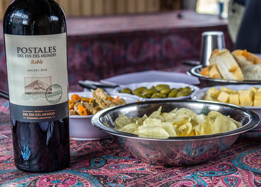 A bottle of Postales del Fin del Mundo Malbec 2018 sits on the table amidst a bowl of chips, plates of bread, olives, cheese, and appetizers, evoking the spirit of adventures to Ushuaia at the end of the world.