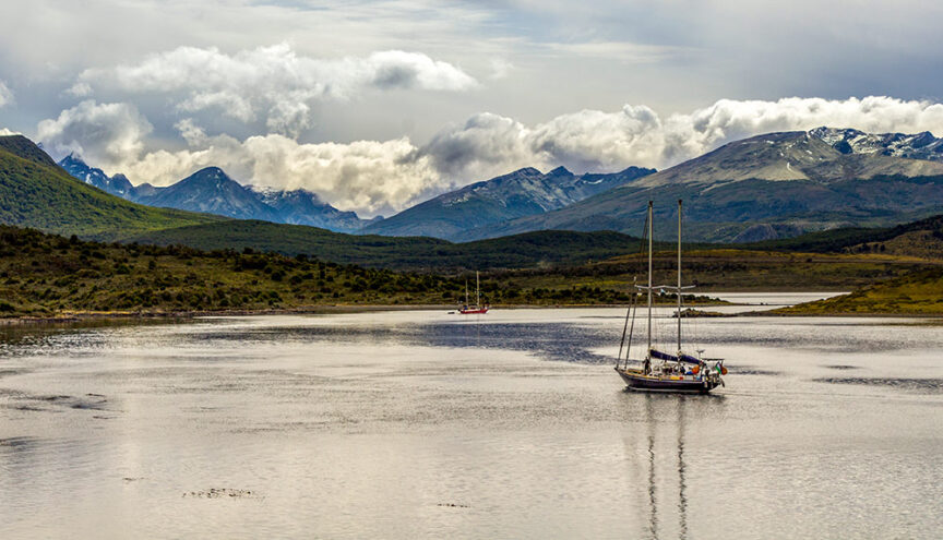 A sailboat floats on a calm lake surrounded by green hills and snow-capped mountain peaks under a cloudy sky, evoking the spirit of adventures. Another sailboat is visible in the distance, reminiscent of the remote waters near Ushuaia.